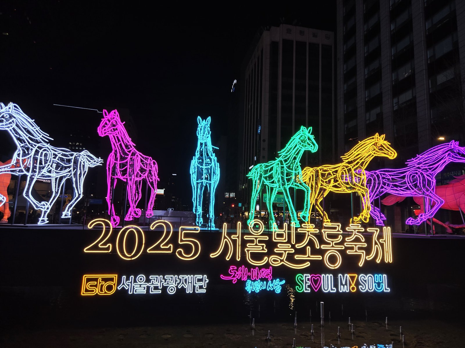 Neon wire-frame horses lit in white, pink, blue, green, yellow and purple at the 2025 Seoul Lantern Festival, city buildings behind them at night